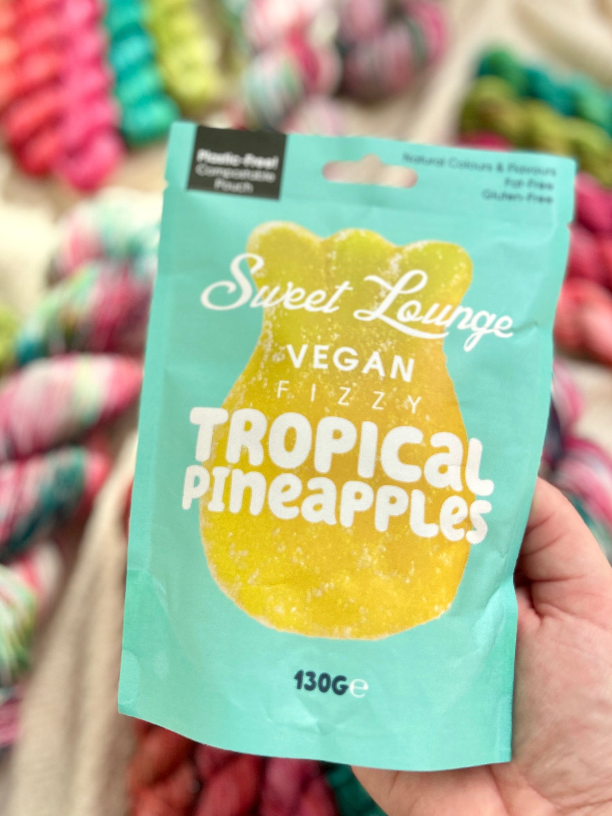 A hand holding a bag of Sweet Lounge Vegan Fizzn Tropical Pineapples candy in front of a blurred background of colorful yarn skeins.